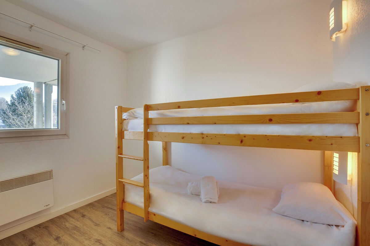 Een stapelbed met houten frame en wit beddengoed, een raam met uitzicht op bomen, en een wandlamp.