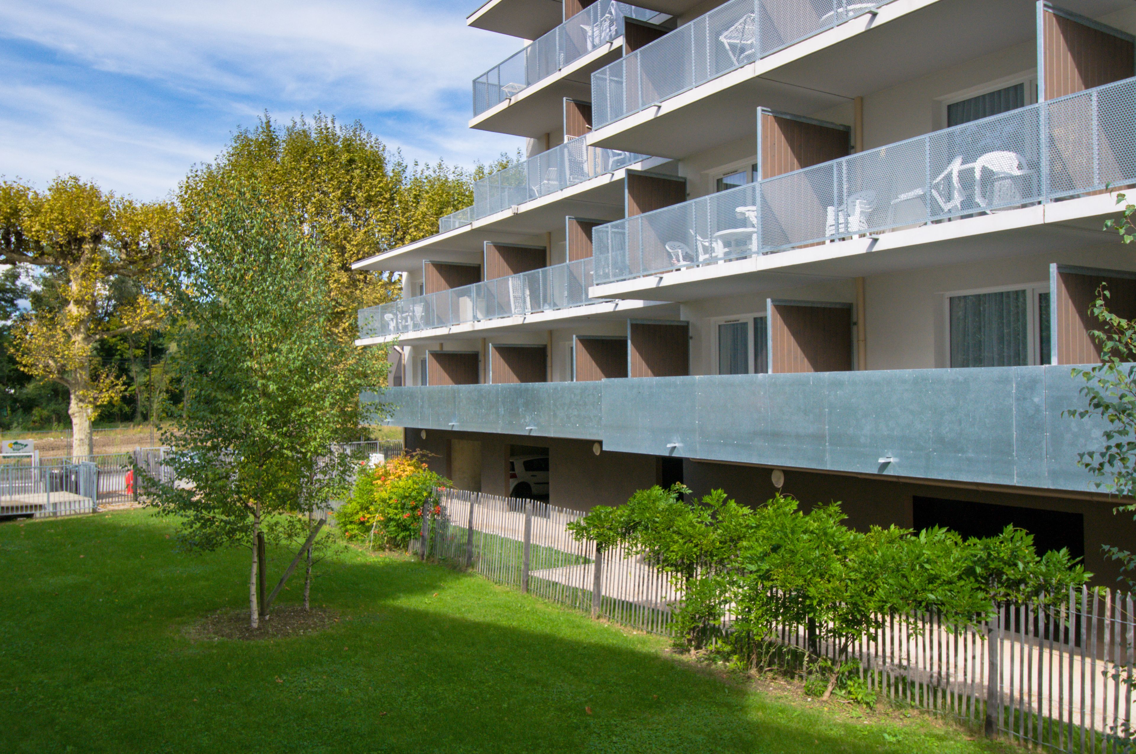 Un immeuble à plusieurs étages doté de balcons, d’un espace de stationnement et d’un petit arbre au premier plan.
