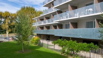 Un edificio de varios pisos con balcones, un estacionamiento y un pequeño árbol al frente.