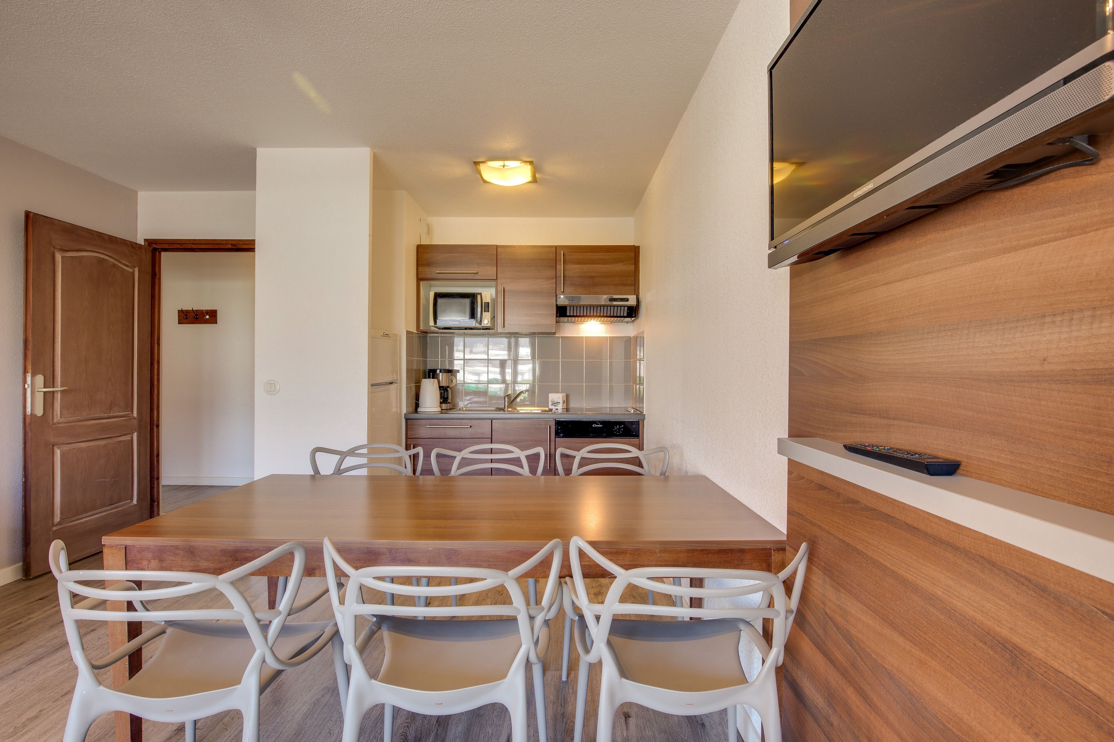 Una cocina moderna con una mesa de madera y sillas blancas, un televisor de pantalla plana montado en la pared y un microondas sobre el fregadero.