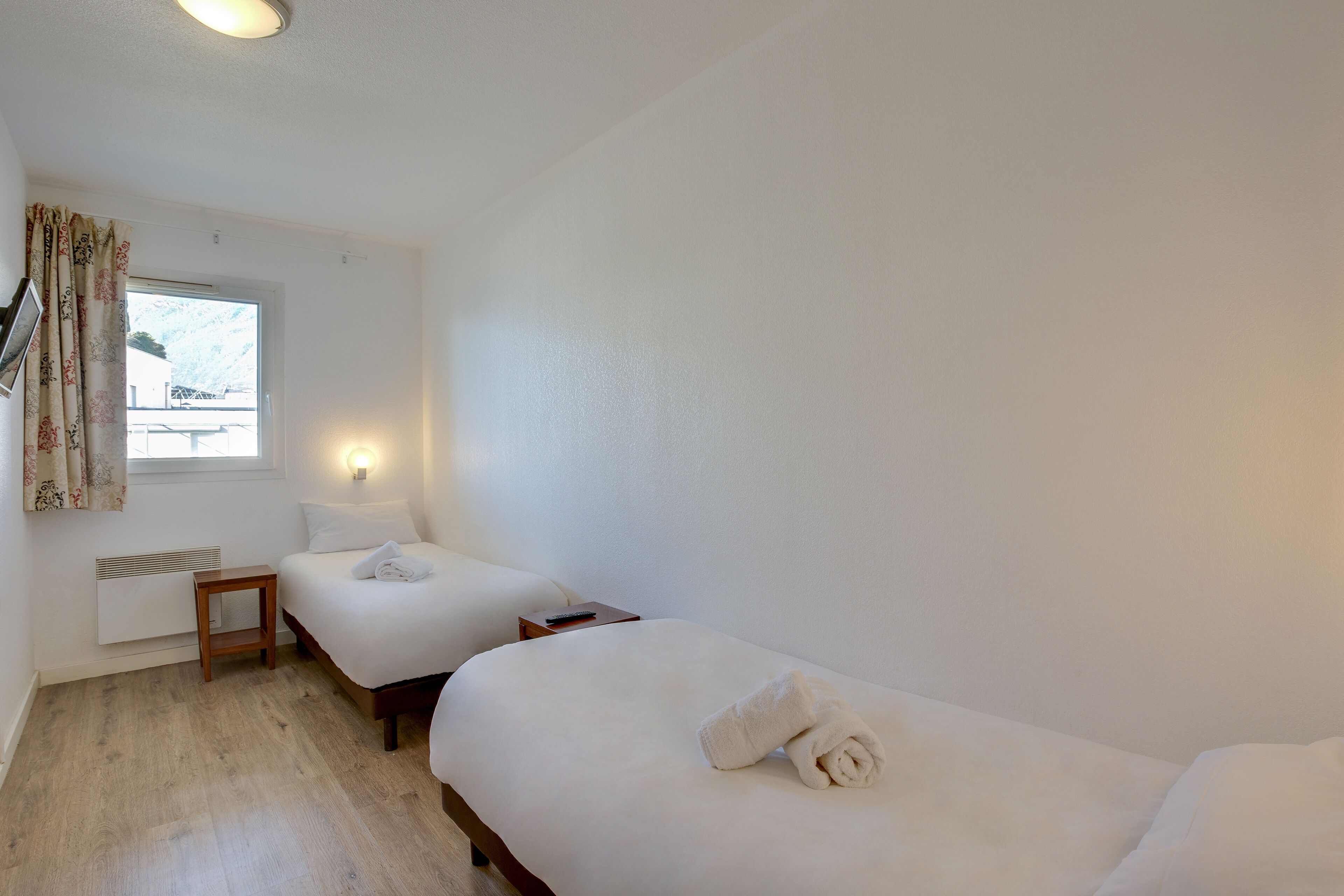 Habitación de hotel con dos camas individuales, una mesita de noche de madera, un calentador fijado a la pared, una ventana con cortinas y una lámpara mural.
