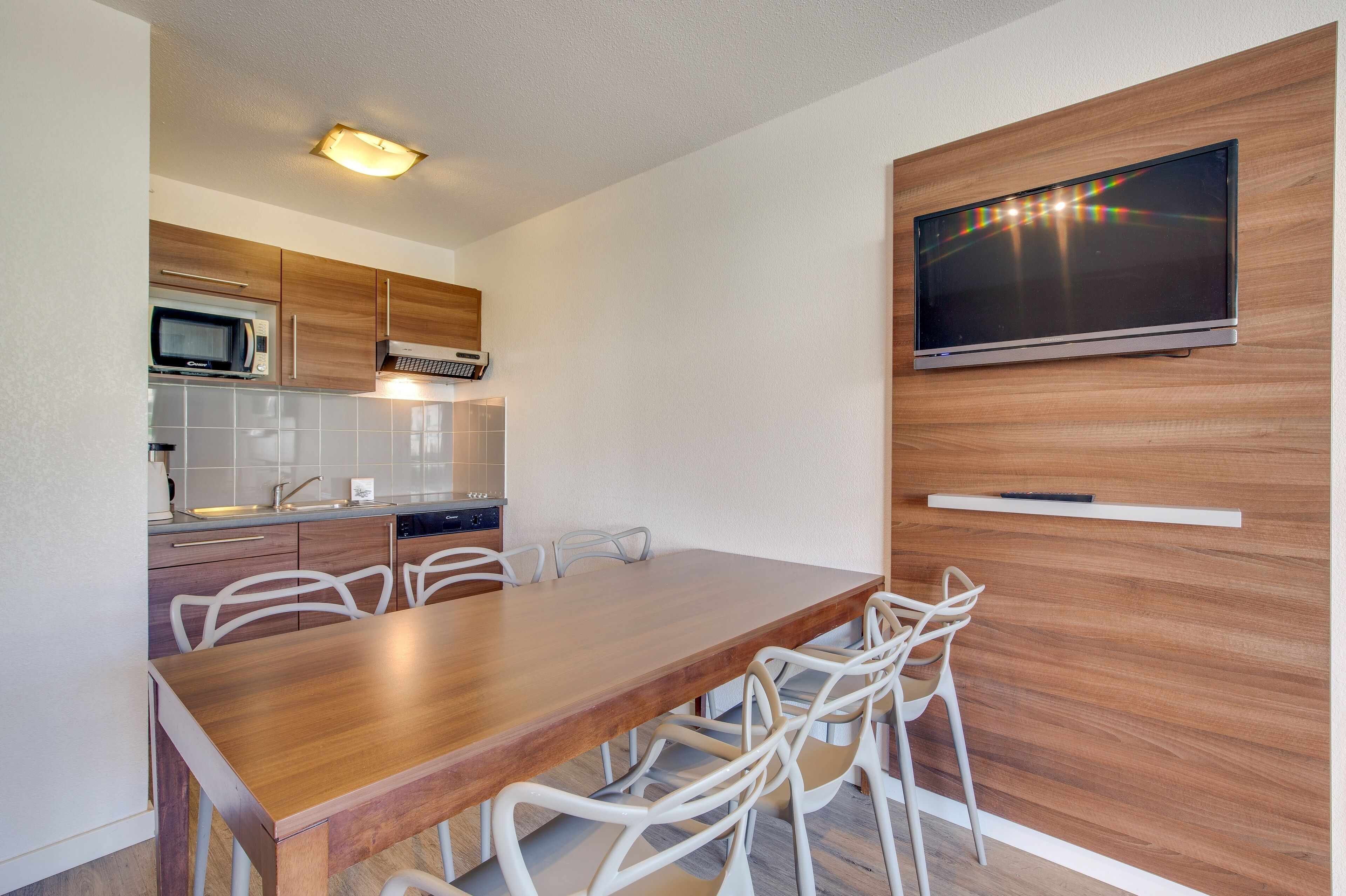 Une cuisine moderne équipée d’un four à micro-ondes, d’une cuisinière et de meubles blancs. On y trouve une table à manger en bois avec des chaises blanches. Un téléviseur à écran plat est fixé au mur, au-dessus de la table.