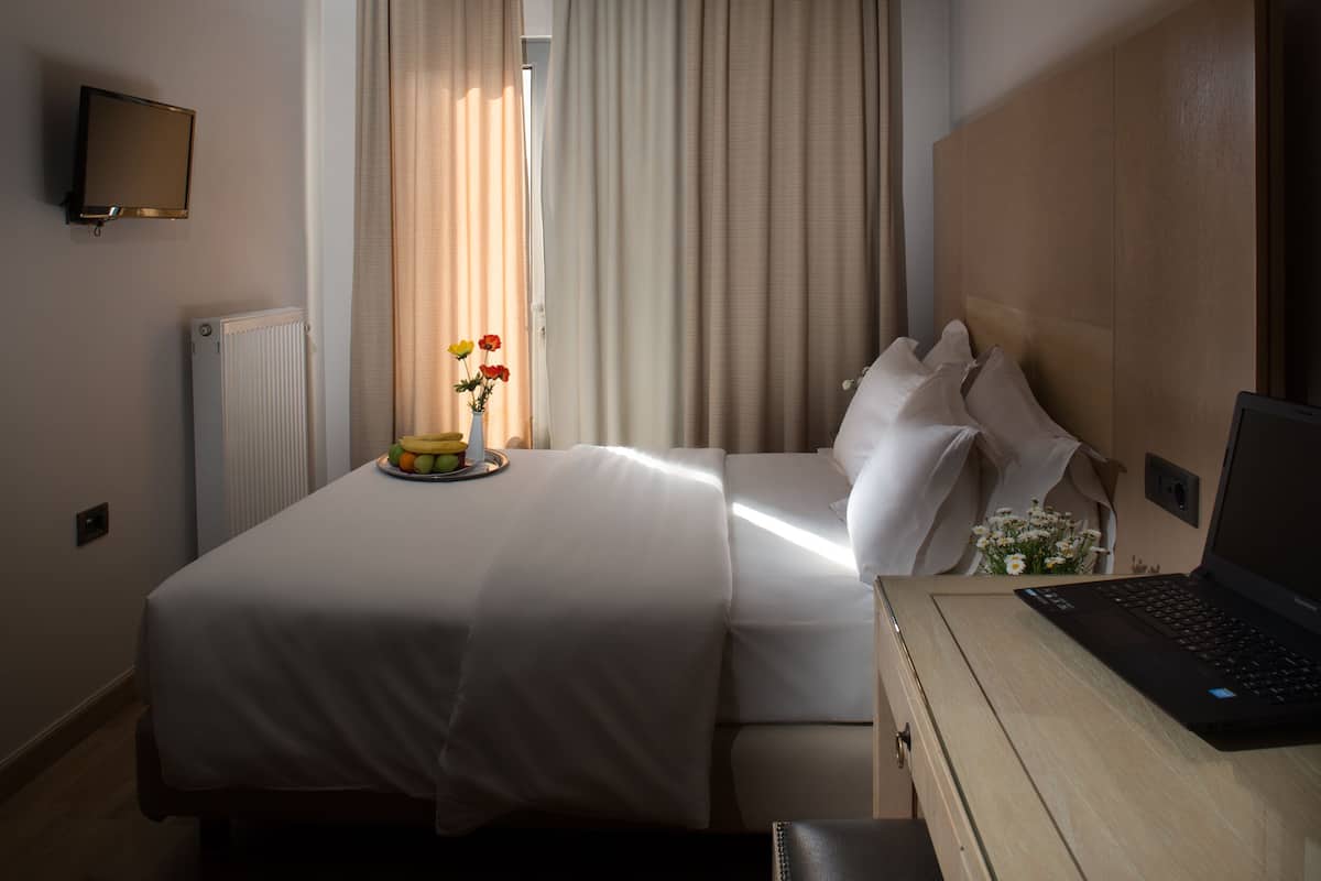 Une chambre d’hôtel avec un lit, une table de chevet sur laquelle se trouve un ordinateur portable, un vase avec des fleurs et un panier de fruits posé sur la table de chevet.