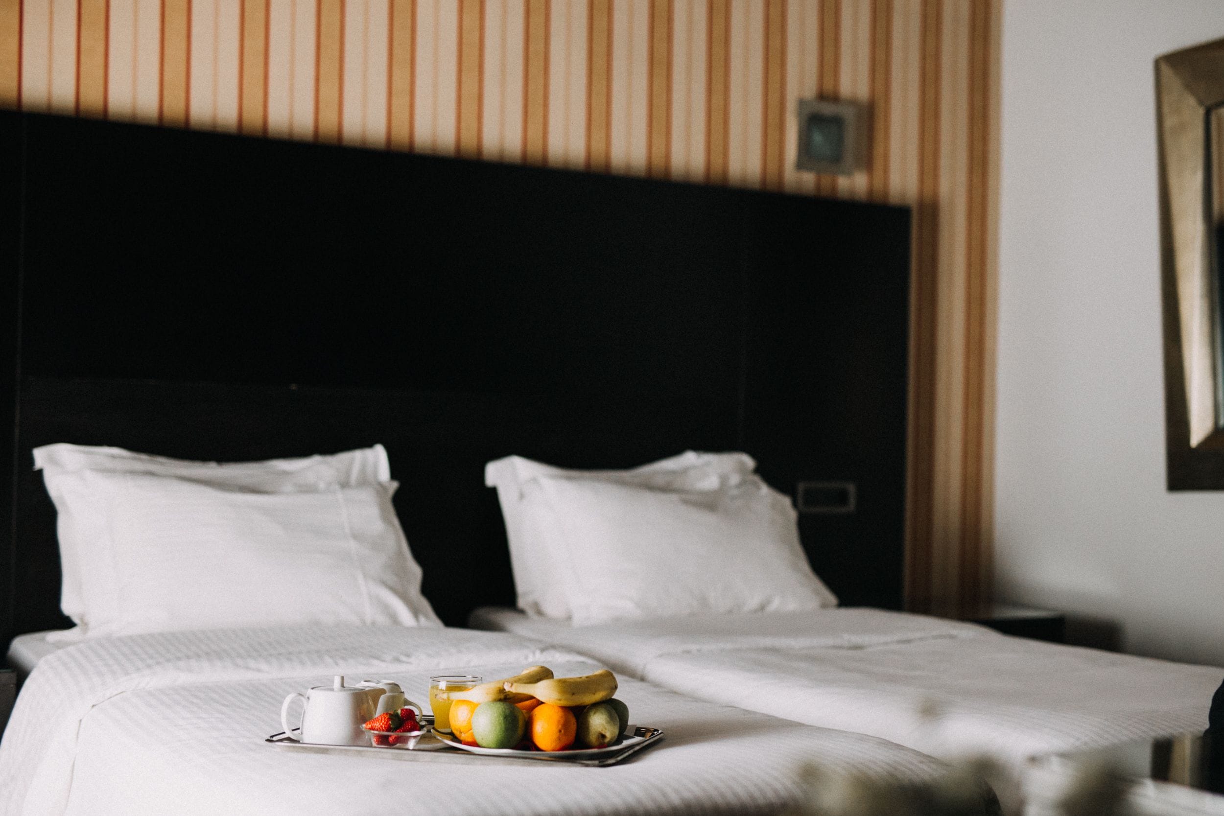 A hotel room with a neatly made bed, a tray of fruit, and a cup of coffee on the bed.