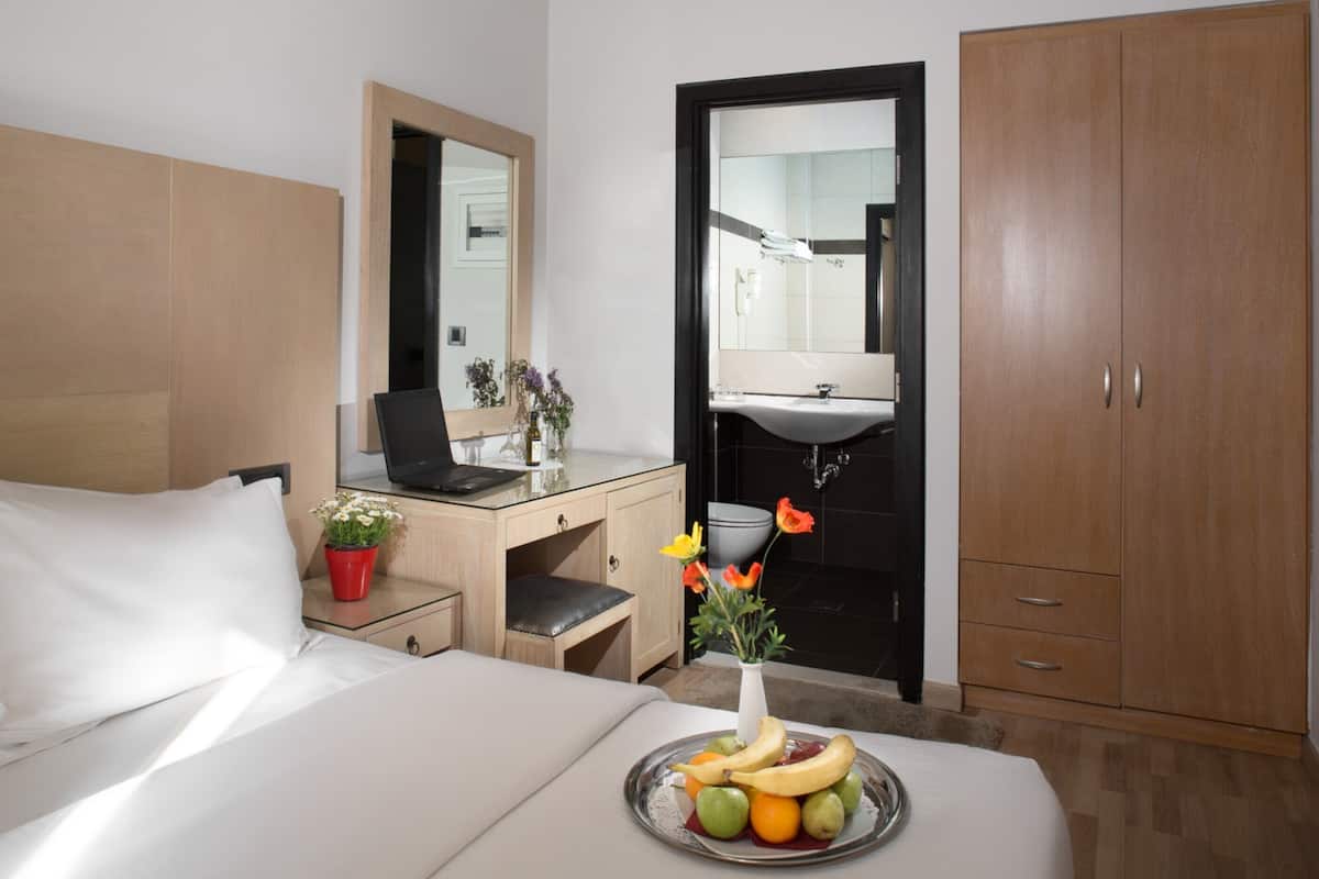 Une chambre d’hôtel avec un lit, un bureau et une chaise, un miroir, une armoire, une salle de bain avec un lavabo et une assiette de fruits sur le lit.