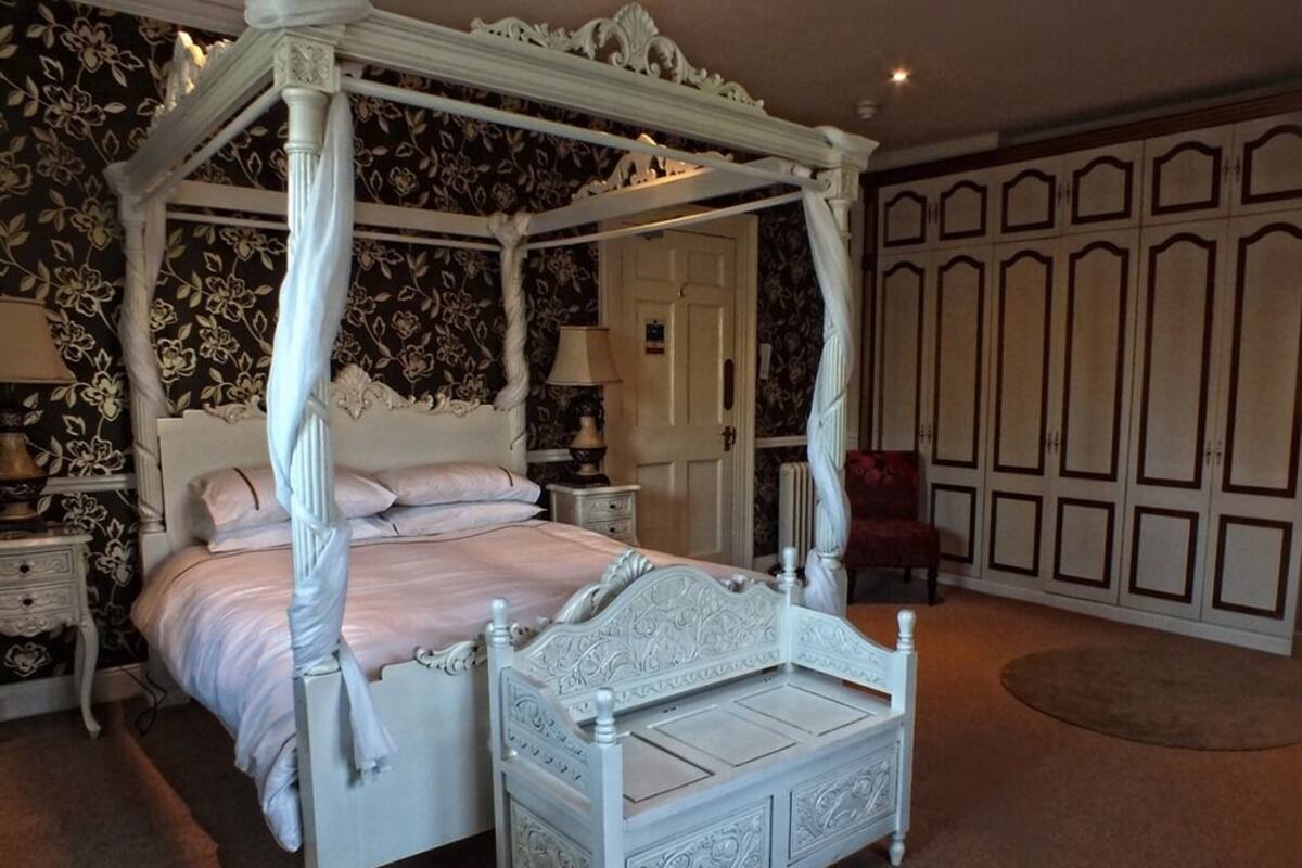 Een bed met hemelbed en vier kolommen, met een witte afwerking, een nachtkastje met lamp, en een rode stoel in een kamer met behang met een patroon en een grote kledingkast.