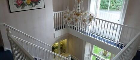A high-ceilinged room with a chandelier, a staircase, and a large window offering a view of greenery.