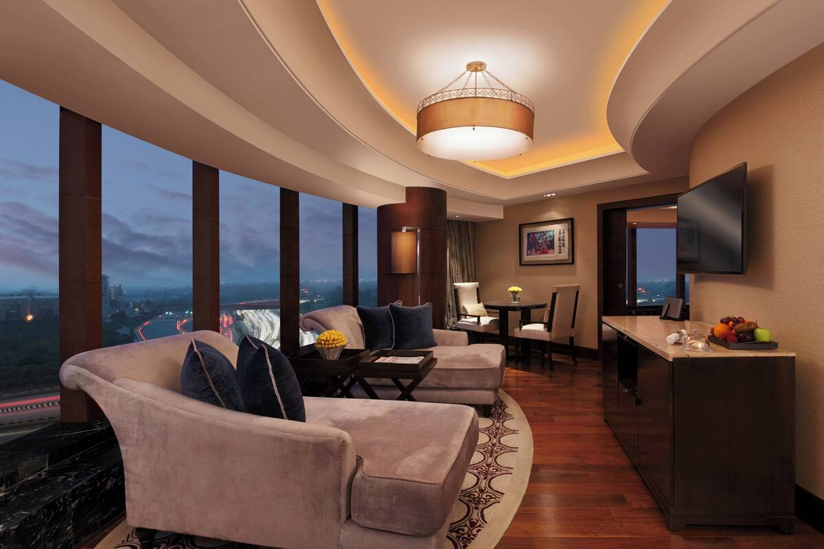 Habitación de hotel moderna con techo curvo, un ventanal grande con vistas a la ciudad, una zona de estar con sofás y una mesa de centro, una mesa de comedor con sillas y un televisor de pantalla plana montado en la pared.