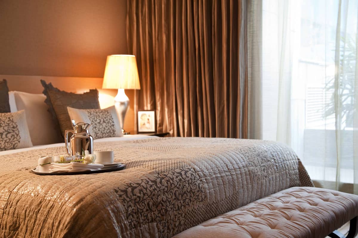 Una cama bien hecha con una bandeja de té y una tetera, una lámpara de noche y una ventana con cortinas.