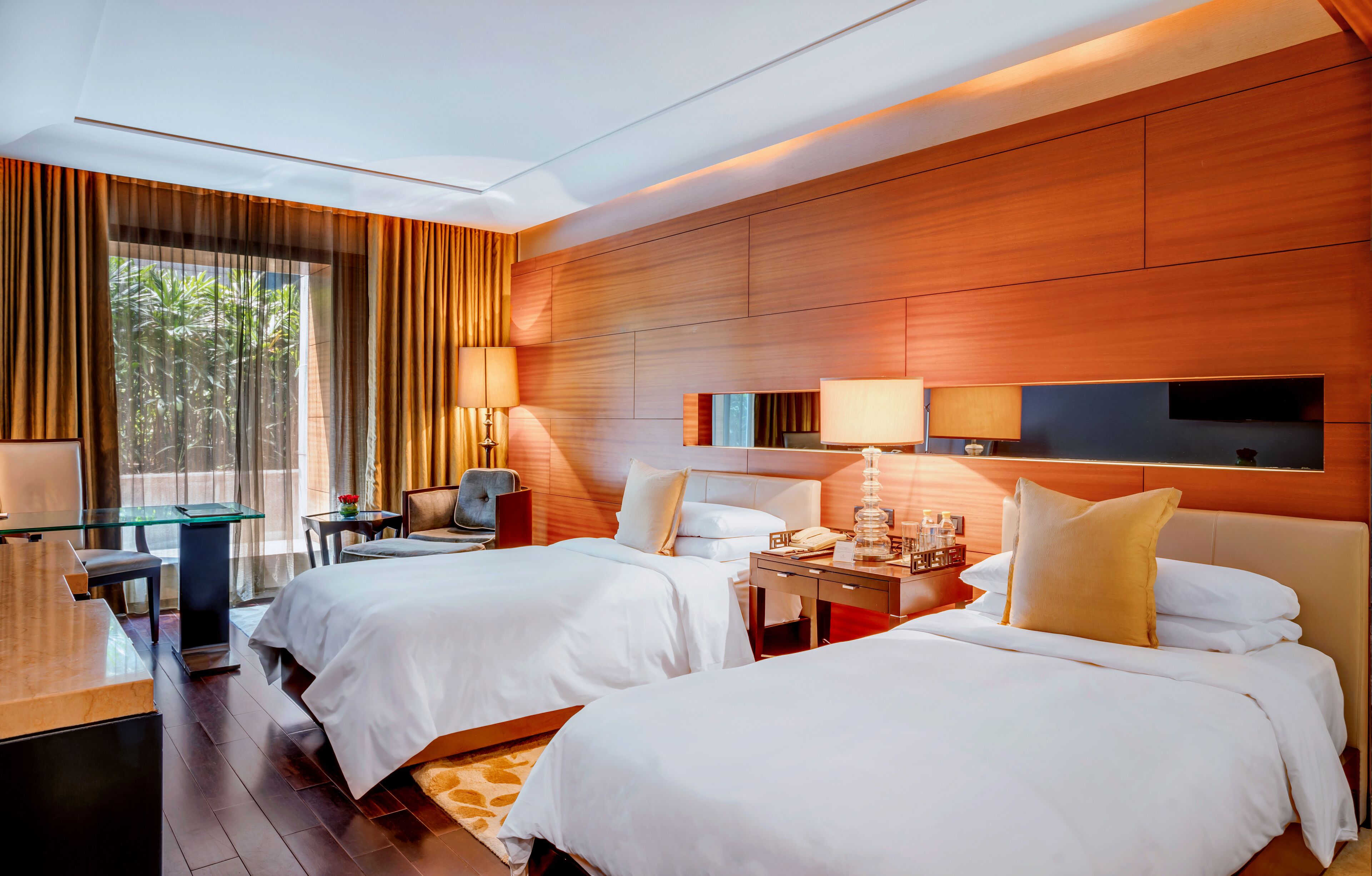 A hotel room with two beds, a desk, a TV, and a large window with curtains.