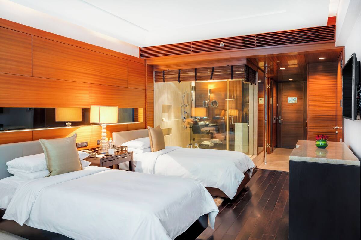 Habitación de hotel con dos camas, un escritorio, un espejo y un baño con cabina de ducha de vidrio.