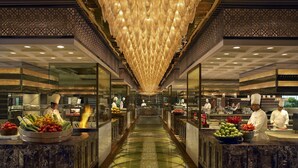A modern kitchen with chefs preparing food, a central glass walkway, and a chandelier. - The Leela Ambience Gurugram Hotel & Residences - The Luxury Urban Sanctuary (Gurugram)