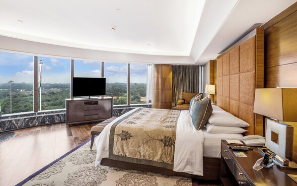 A modern hotel room with a large bed, a flat-screen TV, a desk with a phone and lamp, and a view of the landscape.