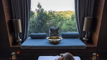 Relax in a serene spa room with a view of lush greenery through large windows, featuring a comfortable seating area with a cushioned bench and decorative bowl.