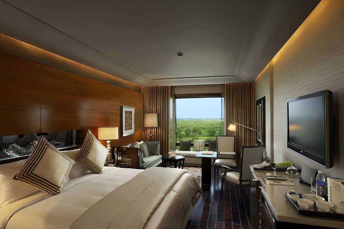 Habitación de hotel con una cama grande, un escritorio con televisor de pantalla plana, una zona de estar con mesa y sillas, y un ventanal con vistas a la vegetación.