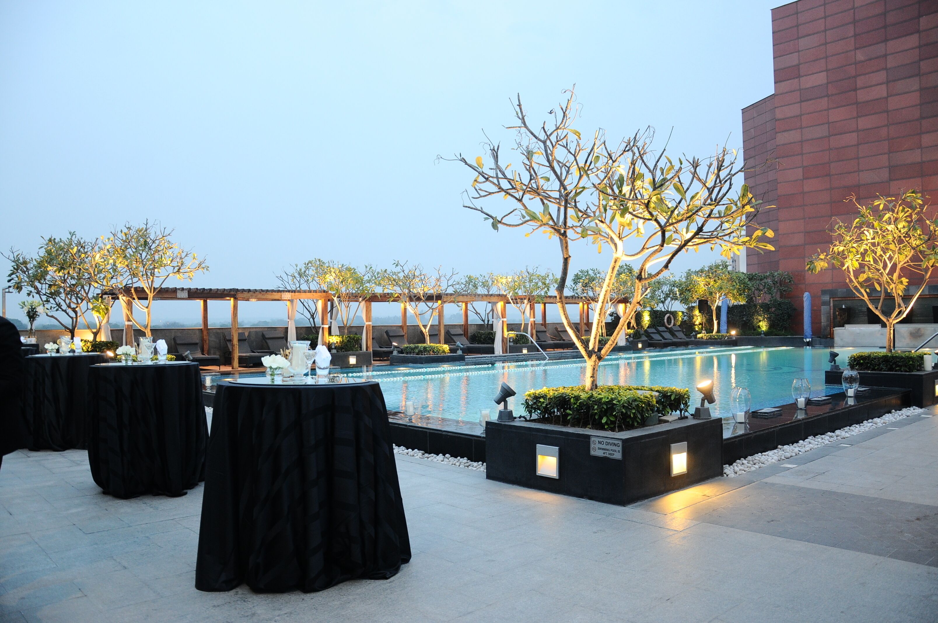 An outdoor pool area with tables set for dining, surrounded by trees and a modern building in the background.