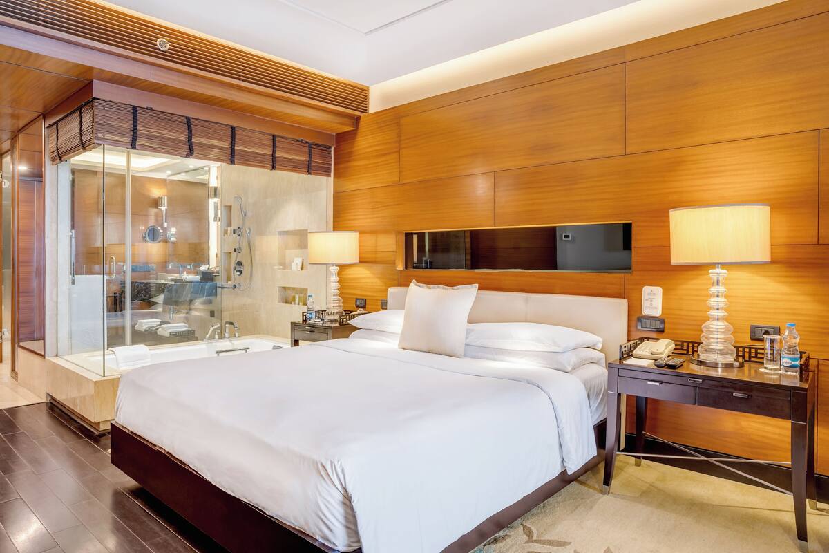 Una habitación de hotel moderna con una cama grande, zona de ducha con paredes de vidrio, paneles de madera y una mesita de noche con lámpara y teléfono.