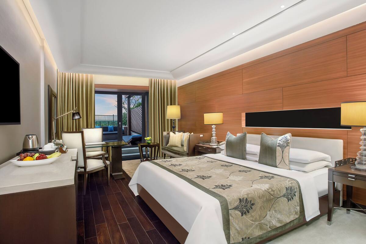 Habitación de hotel con una cama grande, un escritorio con silla, una mesita de noche con lámpara y vistas al exterior.