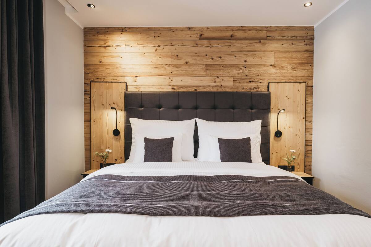 A modern hotel room with a wooden panel wall, a tufted headboard, and a neatly made bed with white linens and a gray throw.