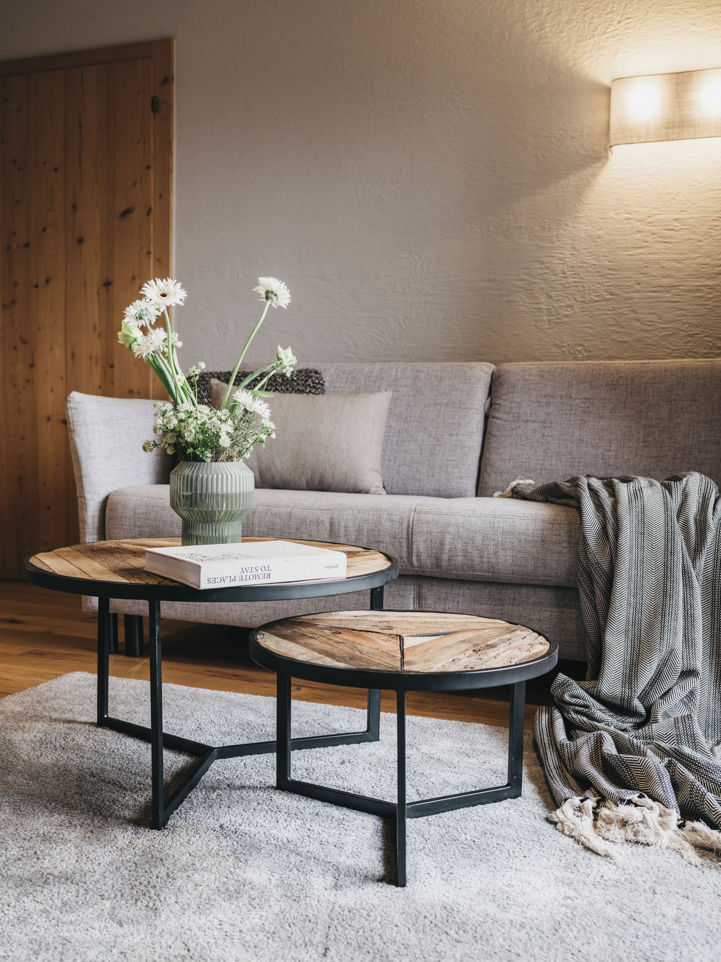 A cozy living room with a grey sofa, two round wooden tables, a vase with flowers, and a lamp.