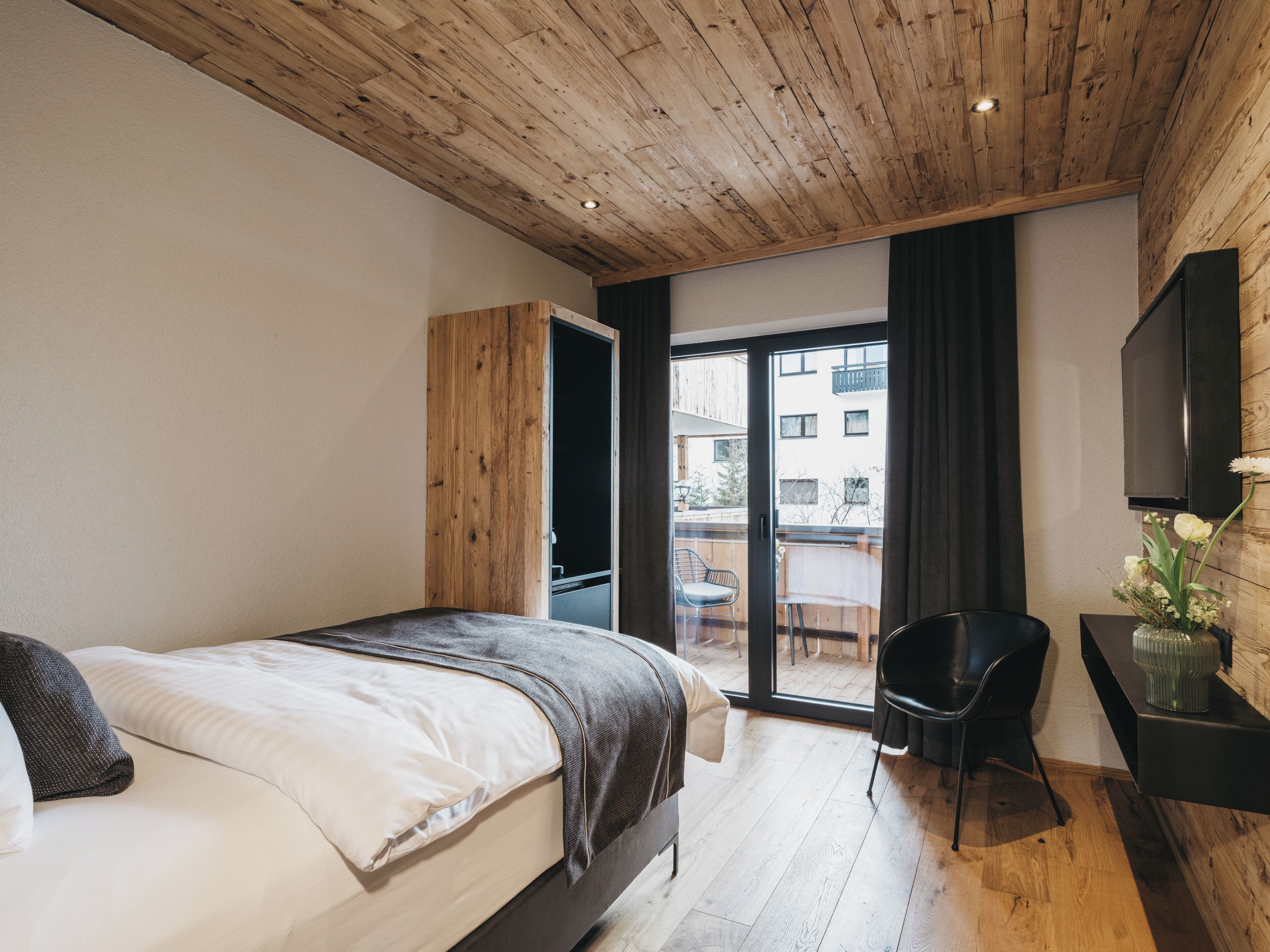 Une chambre moderne avec un plafond en bois, un grand lit, une porte-fenêtre coulissante donnant sur un balcon et un petit bureau avec une chaise.
