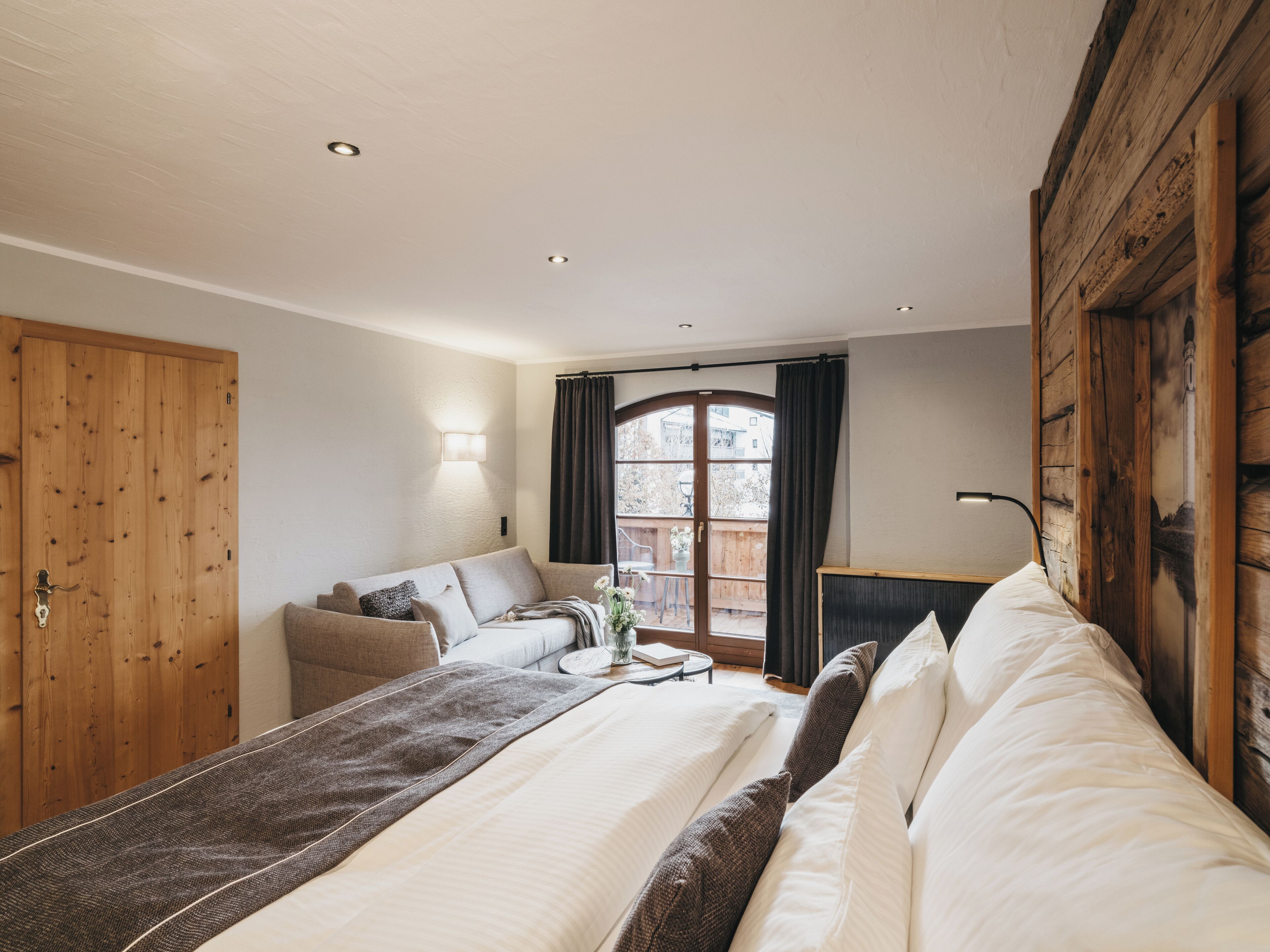 A bedroom with a bed, a sofa, a wooden wardrobe, a window with curtains, and a wall-mounted lamp.
