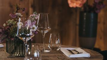 A round wooden table with a vase of flowers, two wine glasses, and a napkin with cutlery.