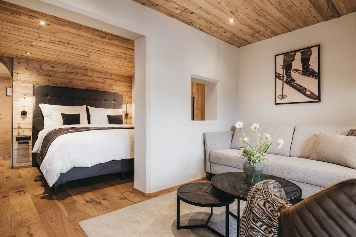 A modern bedroom with a wooden ceiling, a bed with white bedding, a grey sofa, and a small round table with a vase of flowers.
