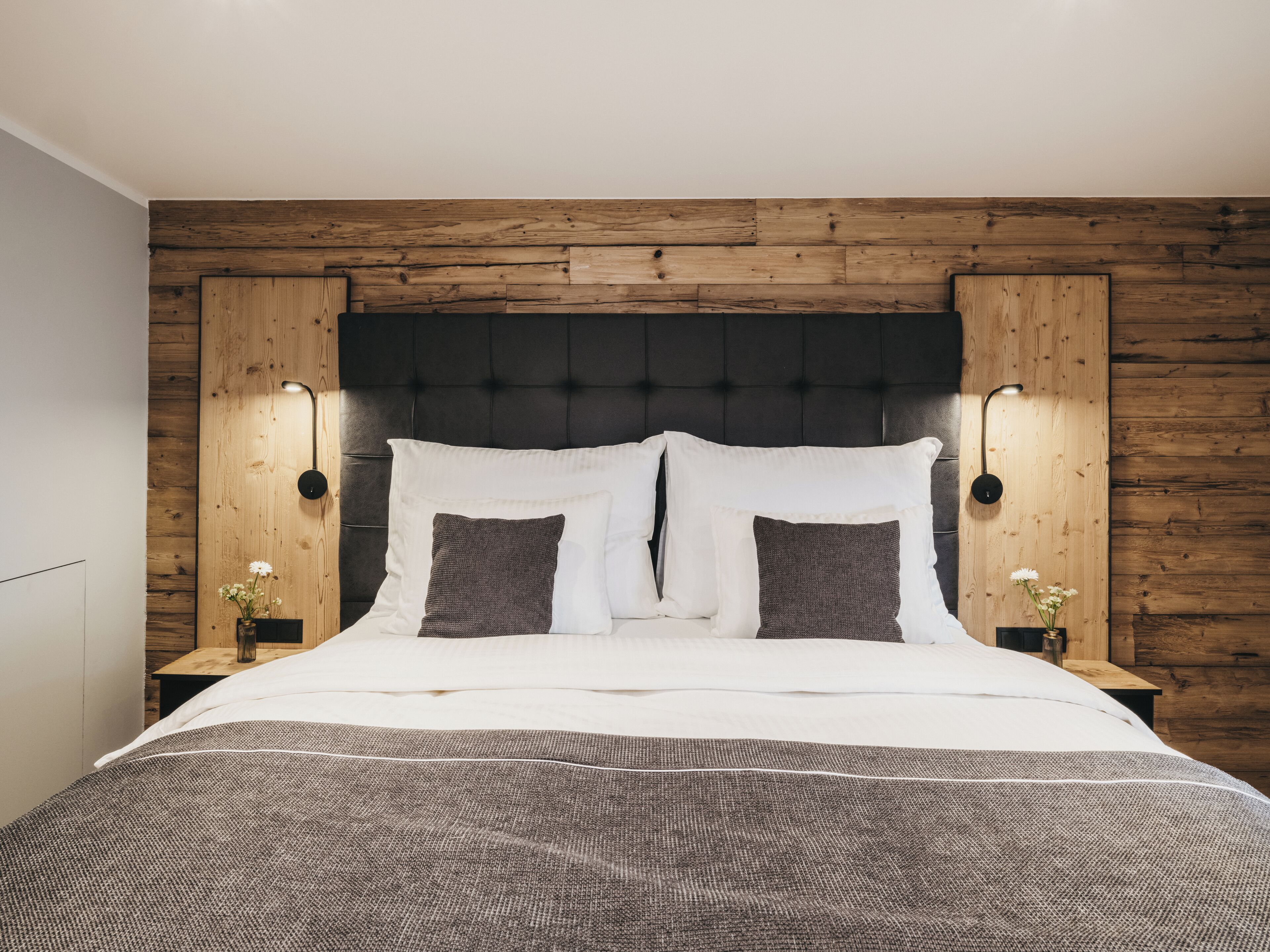 Ein Schlafzimmer mit einem Holz-Kopfteil, zwei Nachttischen mit Lampen und einem Bett mit weißer und grauer Bettwäsche.