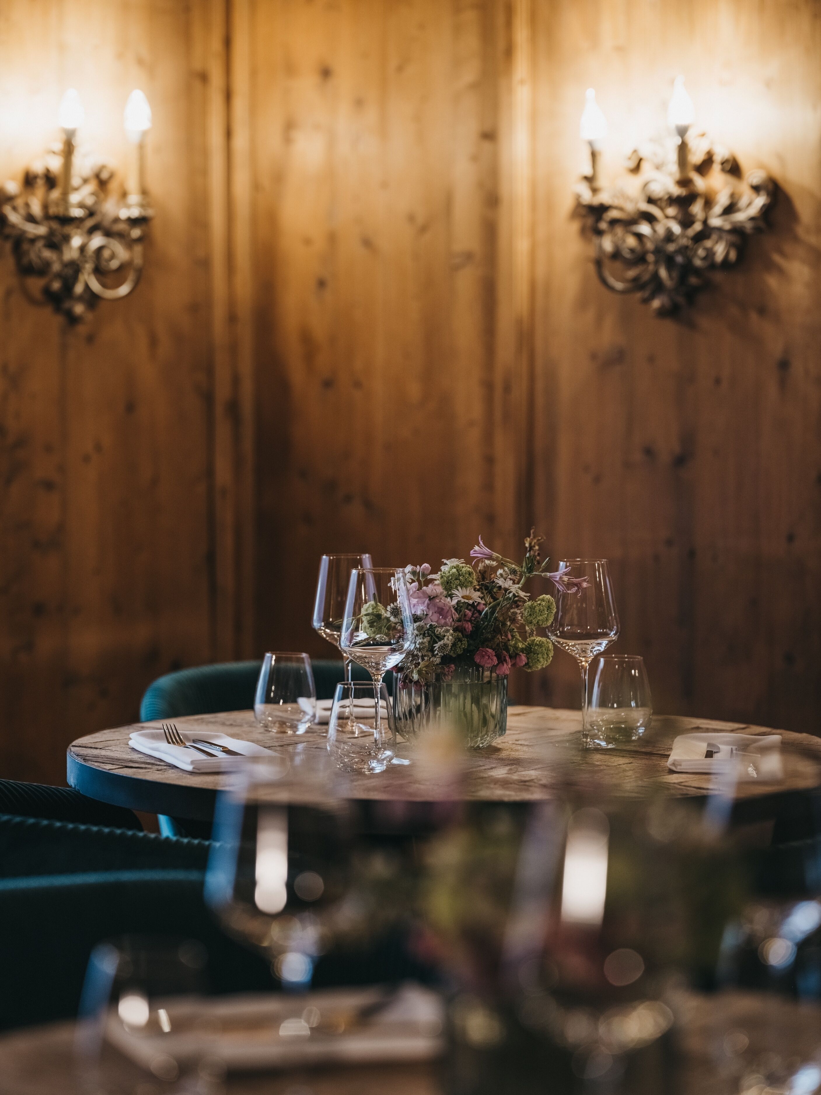 Una mesa redonda para comer con un centro de mesa floral, rodeada de copas de vino vacías y cubiertos, frente a una pared de madera con dos lámparas fijadas.
