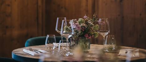 Una mesa redonda para comer con un centro de mesa floral, rodeada de copas de vino vacías y cubiertos, frente a una pared de madera con dos lámparas fijadas.