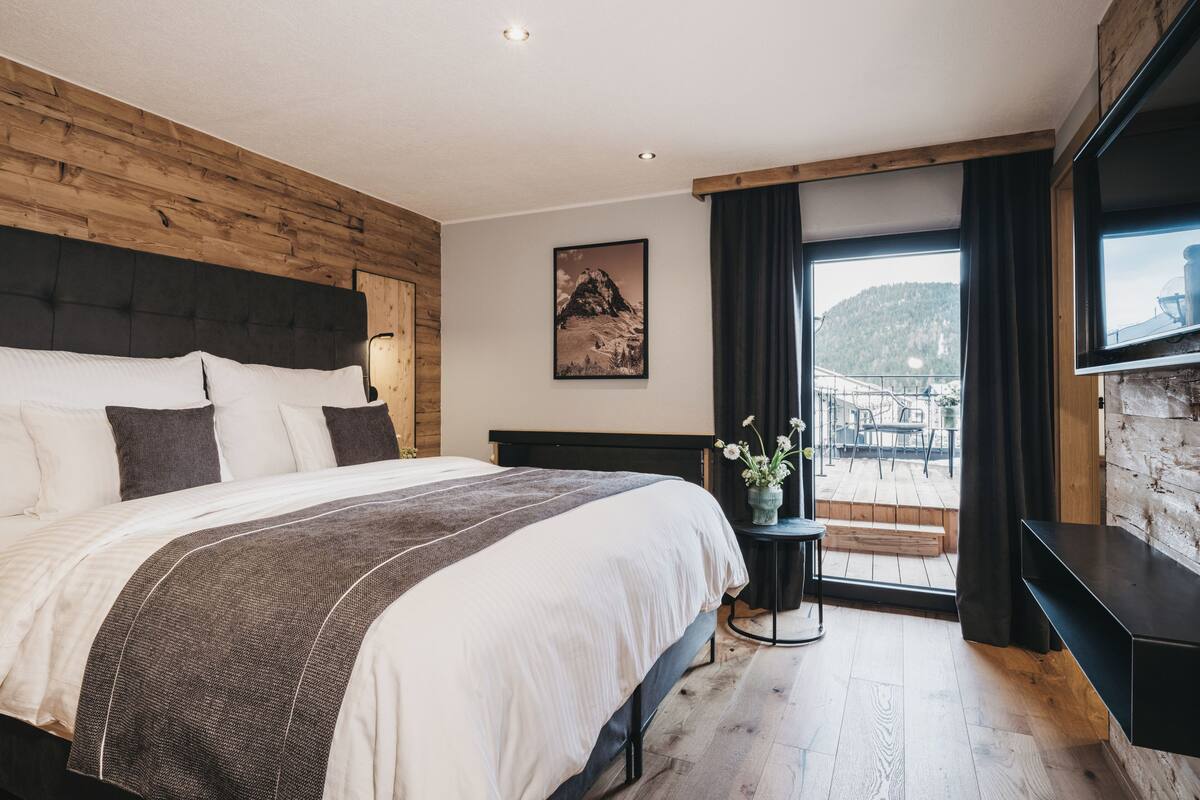 A modern bedroom with a large bed, wooden headboard, a mounted TV, and a balcony with a view of a mountain.
