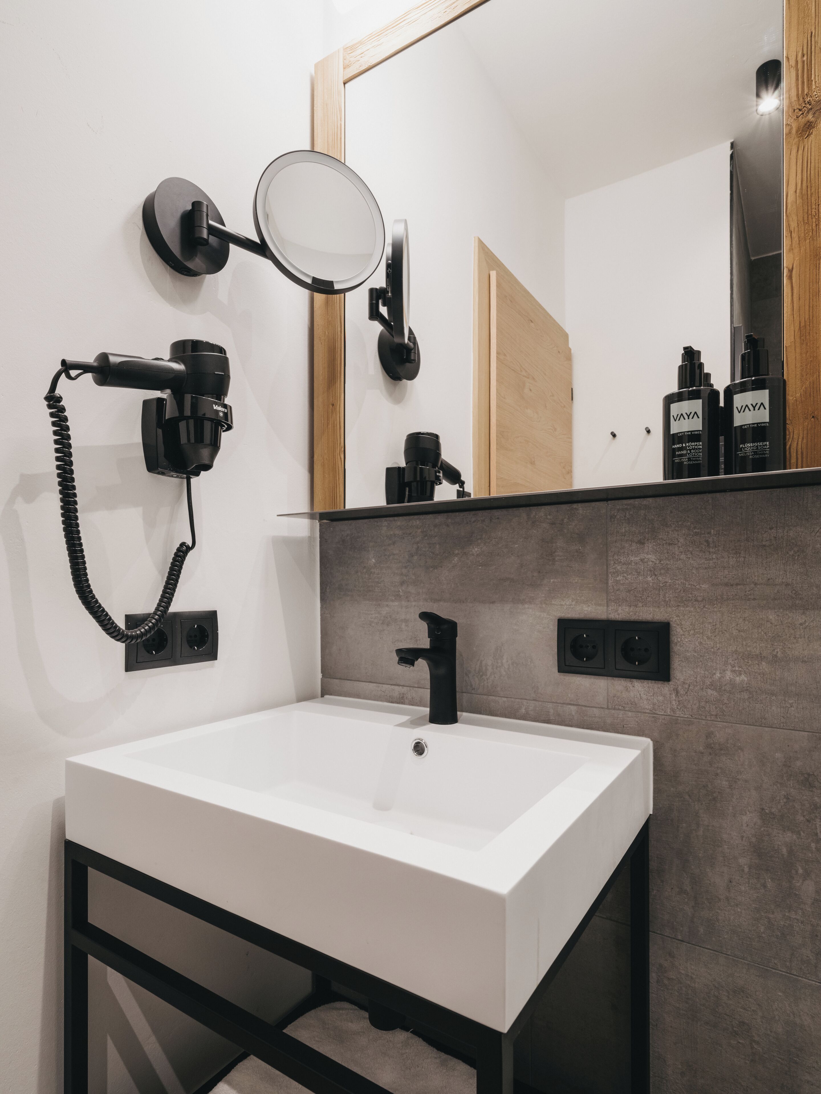 Une salle de bain moderne avec un lavabo blanc, un robinet noir et un miroir rond. On peut voir deux bouteilles noires sur le comptoir, et un sèche-cheveux fixé au mur.