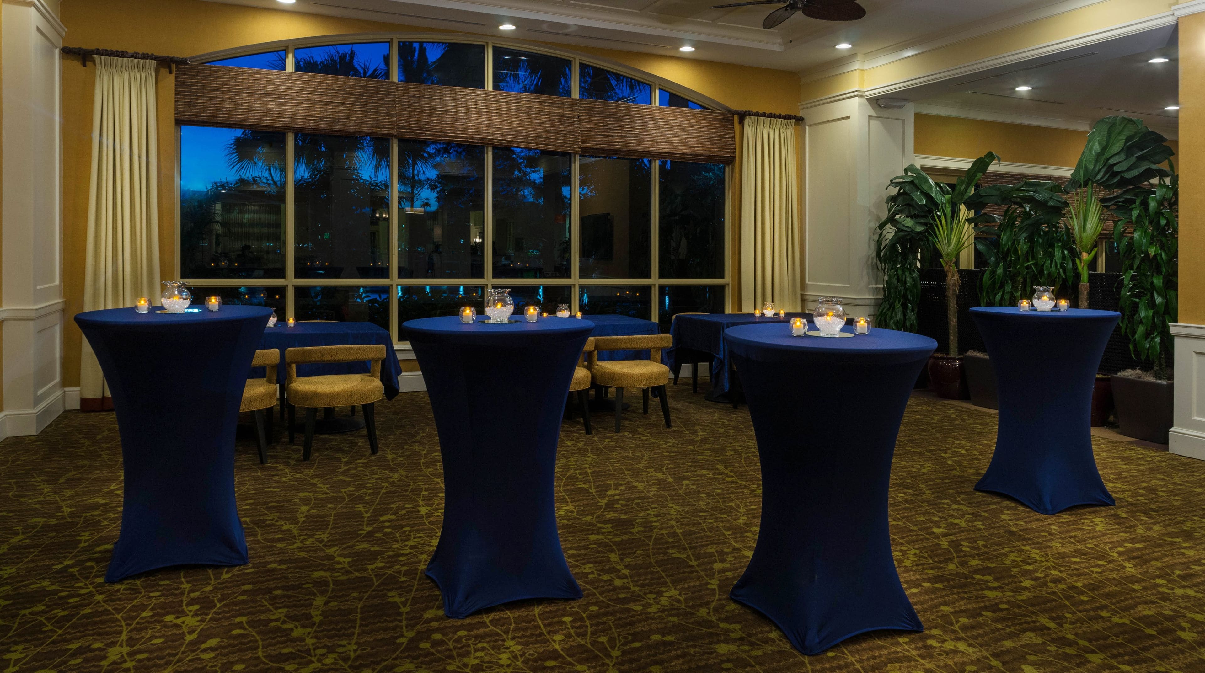 Un salón de conferencias con mesas redondas, sillas y un ventanal con vistas a palmeras.