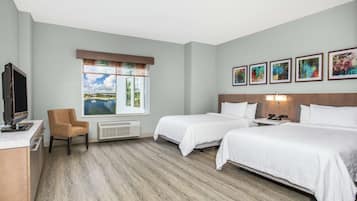 Habitación de hotel con dos camas, un escritorio con televisor, una silla, un ventanal con vistas y varias fotografías enmarcadas en la pared.