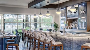A modern bar area with a long counter, multiple stools, and a large window offering a view of the outdoors.