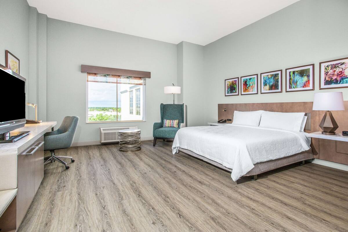 A hotel room with a bed, desk, chair, and artwork on the wall.
