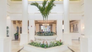 A spacious lobby with a central fountain surrounded by plants, white columns, and a high ceiling.