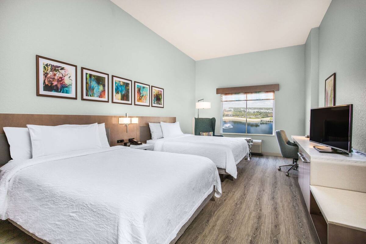 A hotel room with two beds, a desk with a TV, a chair, a lamp, and a window with a view.