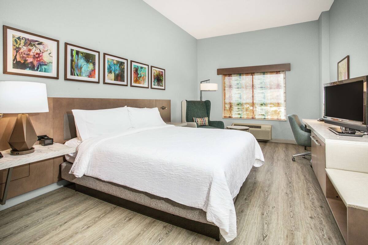 A hotel room with a large bed, a desk with a computer, a chair, and a TV. There are four framed pictures on the wall.