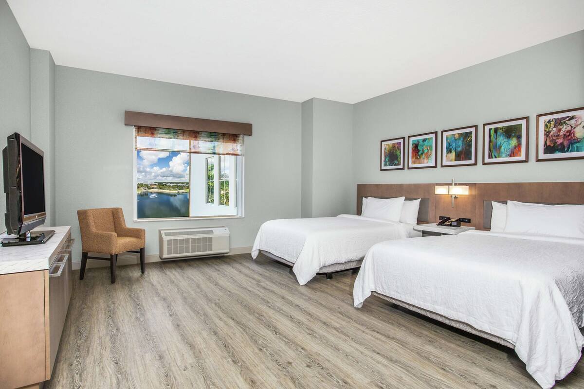 A hotel room with two beds, a desk, a chair, a TV, a window with a view, and artwork on the wall.
