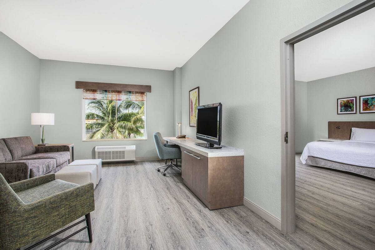 A hotel room with a bed, a desk with a computer, a chair, a couch, and a window with a view of palm trees.