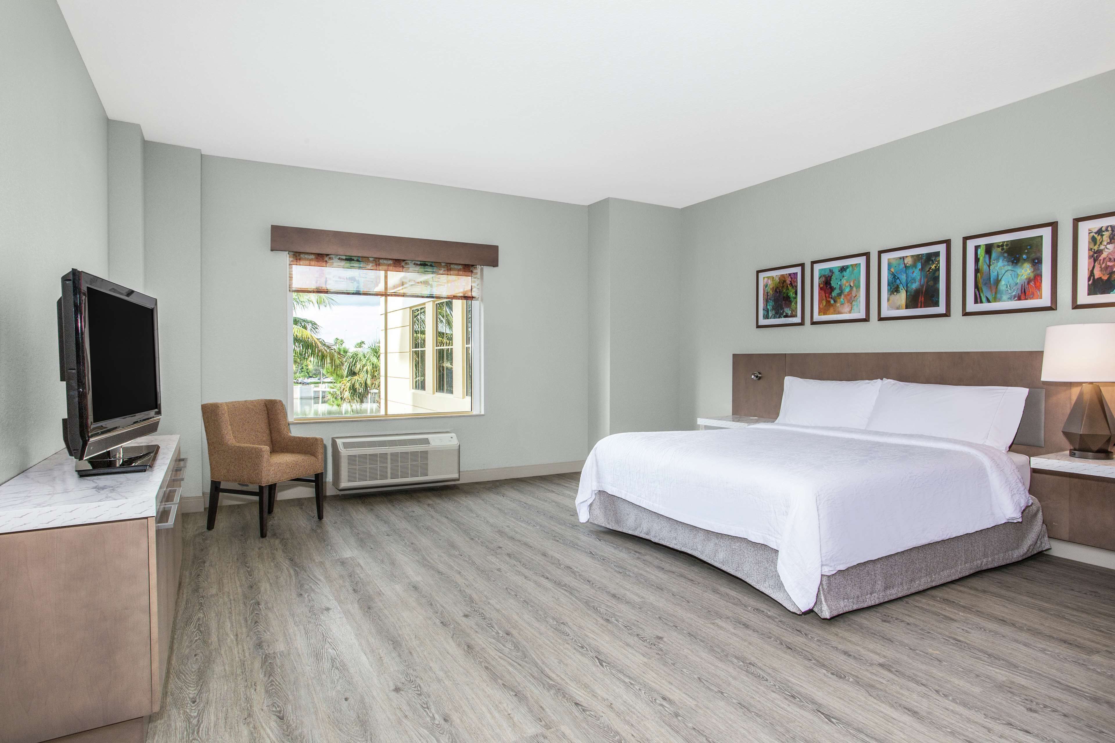 Habitación de hotel con cama, televisión, una silla, ventana con vistas y obras de arte en la pared.