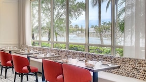 A restaurant with a long counter, red chairs, and a view of palm trees and water.