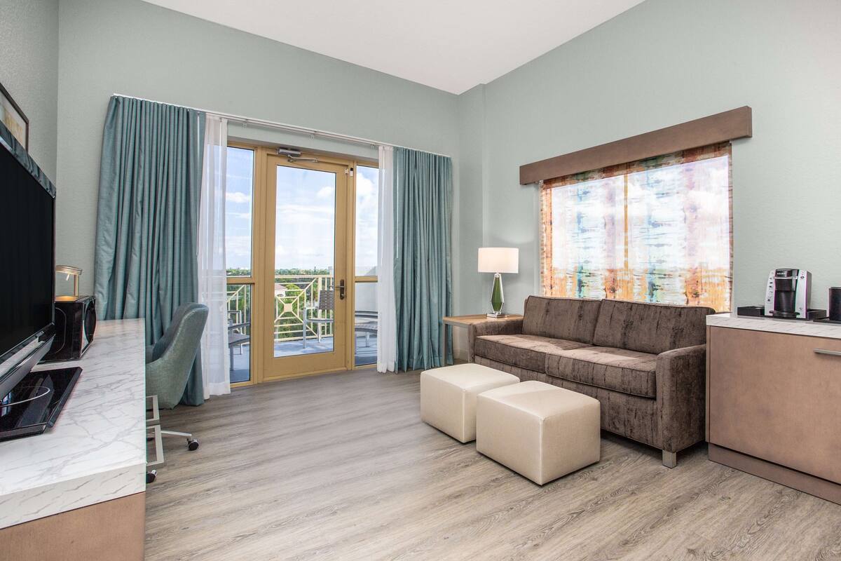 A modern hotel room with a sofa, ottomans, a TV, and a coffee maker.