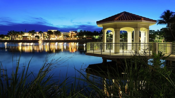 A gazebo with a tiled roof, surrounded by a railing, overlooking a calm body of water with reflections of nearby buildings and palm trees.