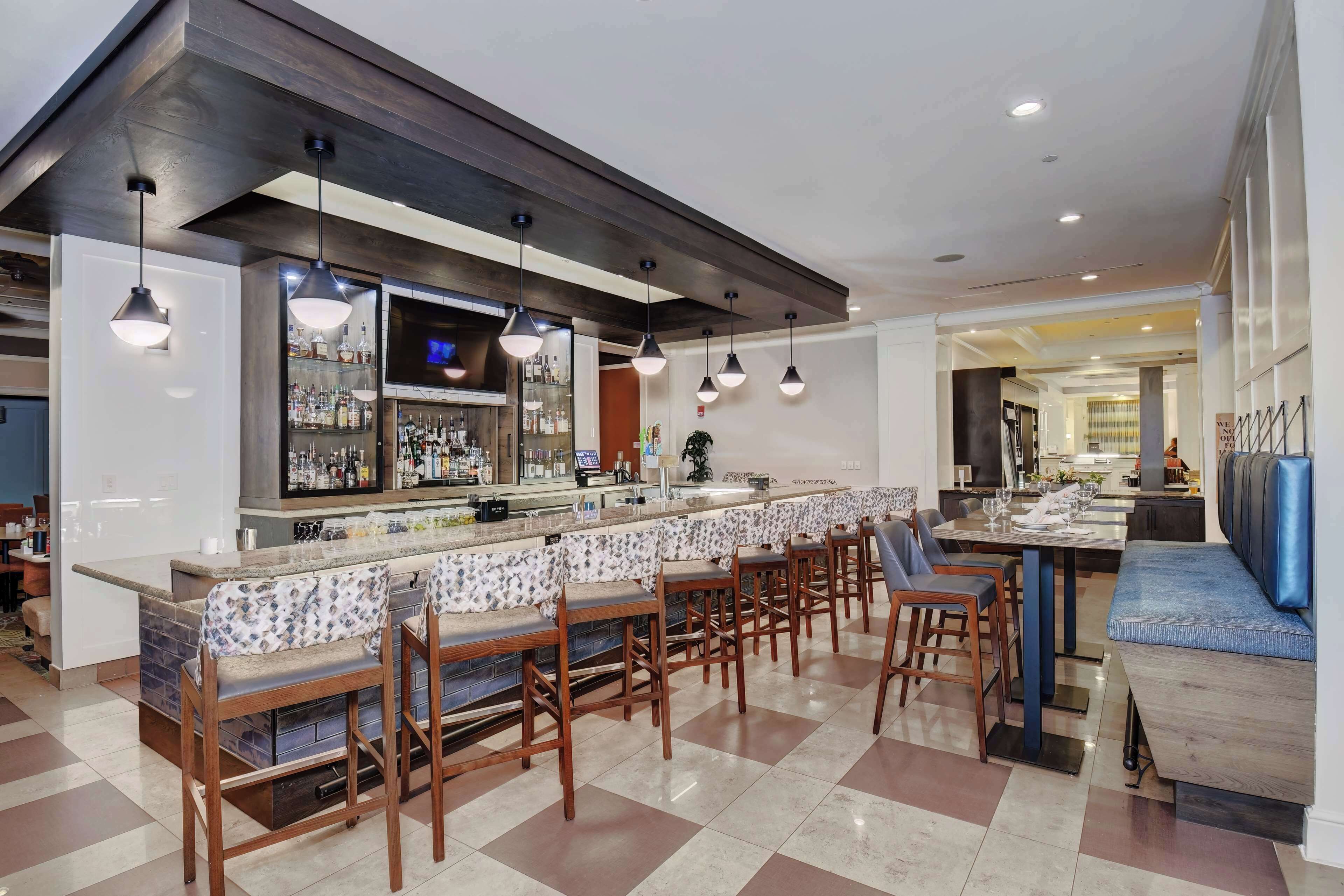 Uma área de bar moderna com um balcão comprido, vários bancos altos e iluminação pendente.