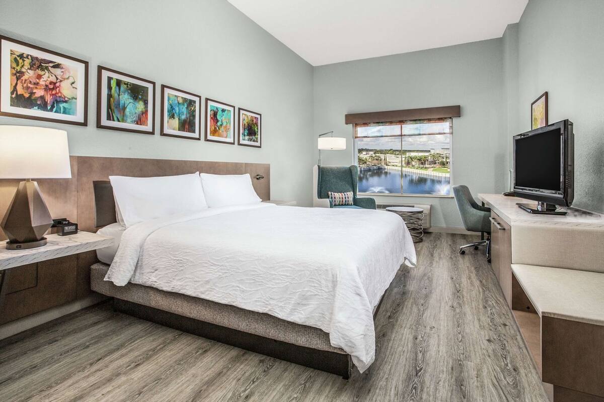 A hotel room with a bed, desk, chair, TV, artwork, and a window with a view.