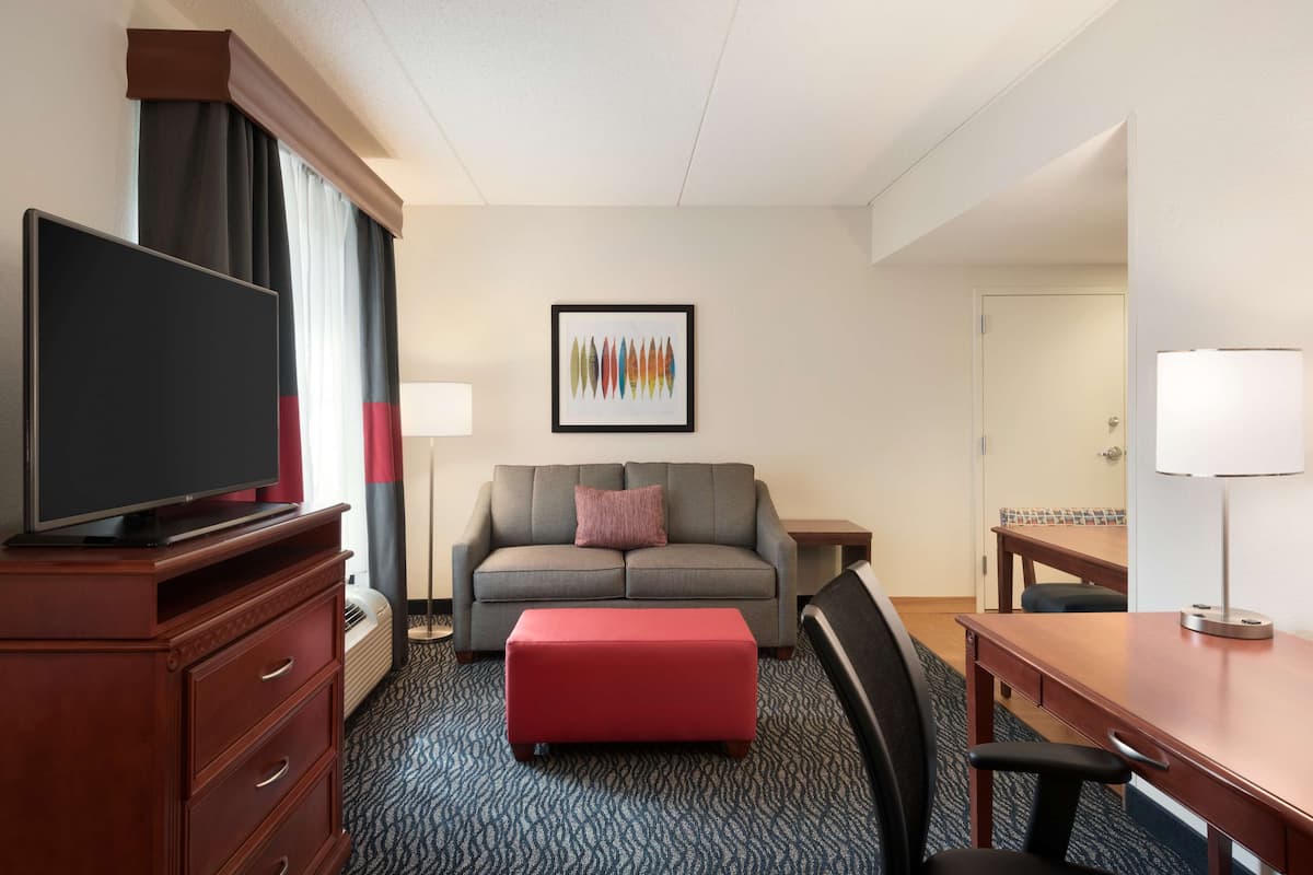 A hotel room with a flat-screen TV, a grey sofa, a red ottoman, a desk, and a framed artwork on the wall.