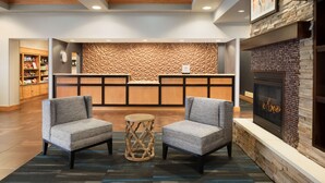 A lobby with two grey armchairs, a round wooden side table, a reception desk with a geometric wall, a fireplace, and a bookshelf.
